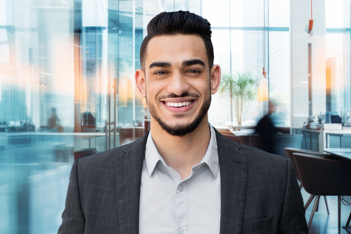 Smiling dark haired male in shirt and blazer in foreground with office setting background