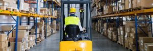 A forklift operator navigates a warehouse aisle, with high shelves stacked with boxes on both sides